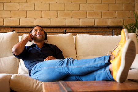 Portrait of cheerful young black man lying on sofa with headphones and listening to musicの写真素材