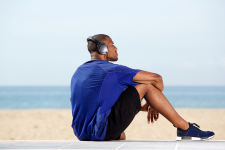 Portrait from behind of fit african man listening to music while sitting outside at the beachの写真素材