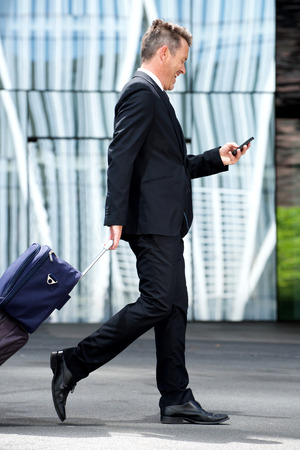 Full length side portrait of business man walking with suitcase and mobile phoneの写真素材