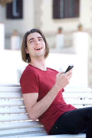 Portrait of young man sitting outside on park bench with smart phoneの写真素材