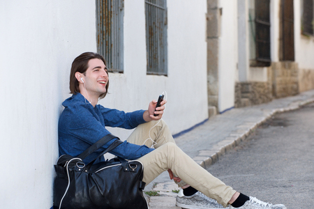 Side portrait of laughing man on city street with smart phone and bagの写真素材