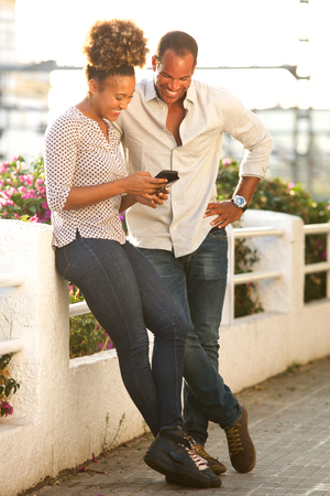 Full length portrait of happy couple standing outside looking at mobile phoneの写真素材