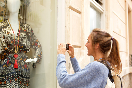 Portrait of happy young woman taking photo of clothes in shop front windowの写真素材