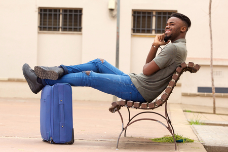 Side portrait of happy african american man sitting outside with suitcase and talking on cell phoneの写真素材