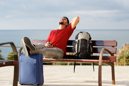 Portrait of happy young man sitting outside with luggage and listening to music with mobile phone and earphonesの写真素材
