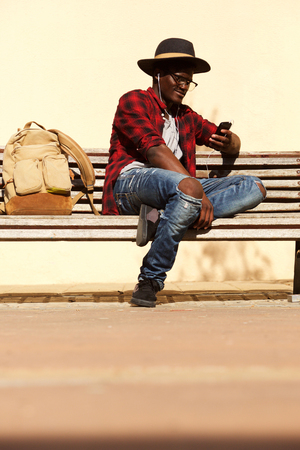 Portrait of stylish young african man sitting on bench using smart phoneの写真素材
