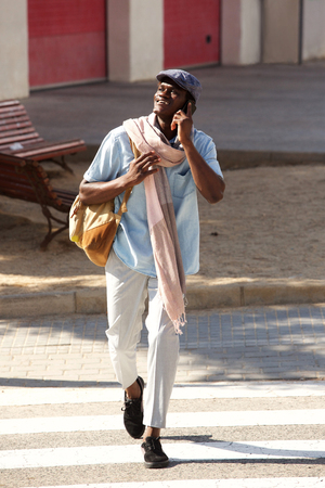 Full length portrait of handsome african man walking outside in the city talking on phoneの写真素材