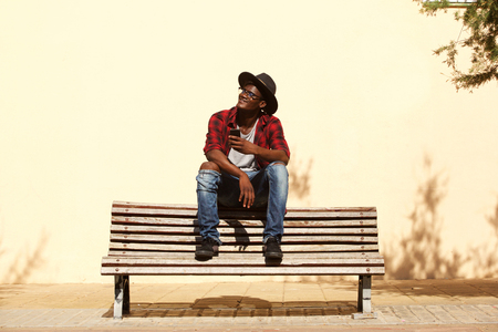 Portrait of cool african guy in hat sitting on bench by the streetの写真素材