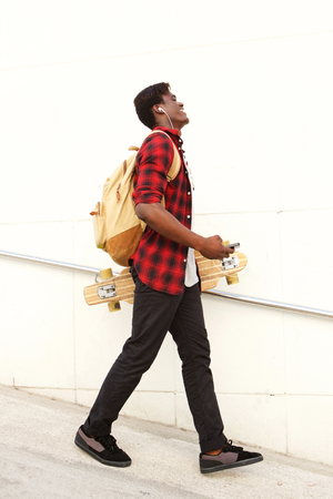 Full length side portrait of handsome young african american male student with skateboard and bag walking outdoorsの写真素材