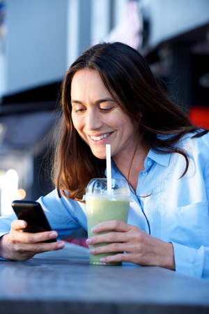 Portrait of happy woman sitting outside with juice and looking at mobile phoneの写真素材