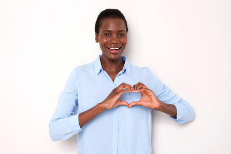 Portrait of beautiful young african woman smiling with heart shaped hand sign on white backgroundの写真素材