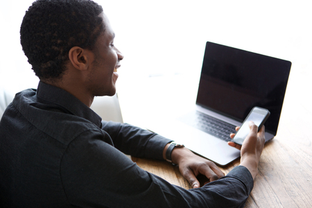 Portrait of young african american man sitting at desk with laptop sending text with cellphoneの写真素材