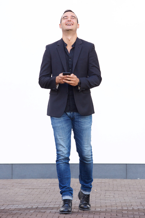 Full body portrait of happy man walking with cellphone by white wallの写真素材