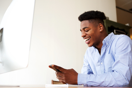 Portrait of happy young african american man sitting with mobile phone at computerの写真素材