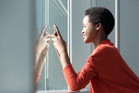 Profile portrait of beautiful smiling african american woman looking at mobile phone by window