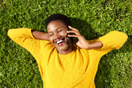 Portrait from above of happy young black woman lying on green grass and talking with mobile phoneの写真素材