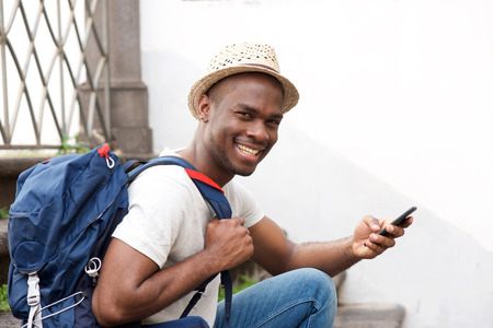 Side portrait of happy african american male tourist sitting on stairs with bag and cellphoneの写真素材