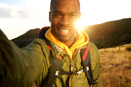 Portrait of happy black man hiking and taking selfie with sunset in backgroundの写真素材