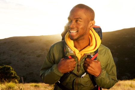 Close up portrait of handsome young african american man with backpack smiling with sunrise in backgroundの写真素材