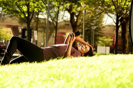 Portrait of happy african american guy relaxing in park and looking at cellphone