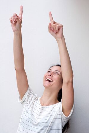 Portrait of cheerful young asian woman laughing with arms raised and fingers pointing up against white wallの写真素材