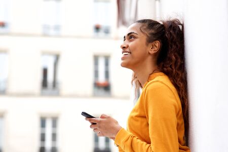 Side portrait of happy young indian woman leaning against white wall and holding mobile phone