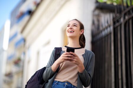 Portrait of smiling young woman with bag holding mobile phone and looking away