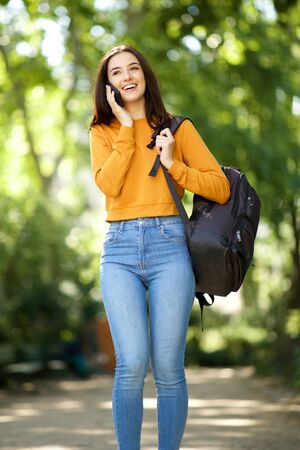 Portrait of female college student walking through park with mobile phone and bag