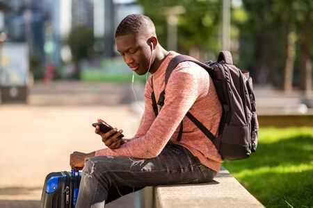 Portrait of young black travel man sitting outside with mobile phone and bagsの写真素材
