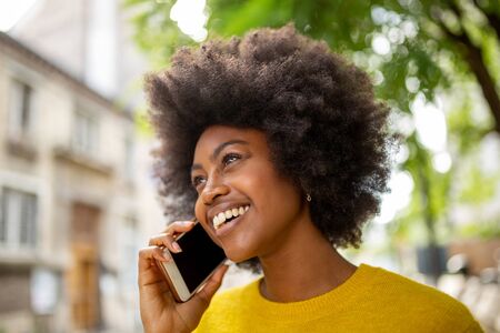 Close up portrait of attractive young african woman smiling and talking with cellphone outsideの写真素材