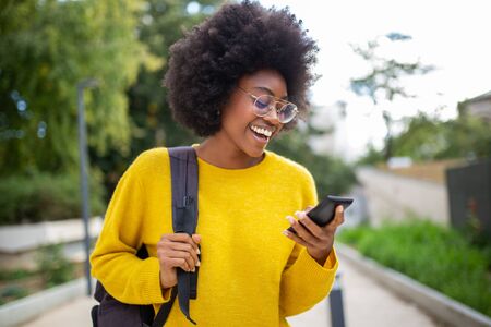 Portrait of smiling young black woman with glasses and bag looking at cellphone outdoorsの写真素材