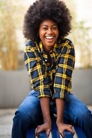 Portrait of happy stylish young black woman with afro hair sitting outside on benchの写真素材