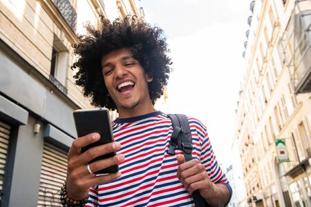 Close up portrait of happy young arab guy with afro hair looking at cellphone in city and laughing