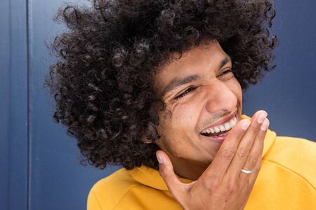 Portrait of young North African man with afro hair laughing with hand covering mouth by blue backgroundの写真素材