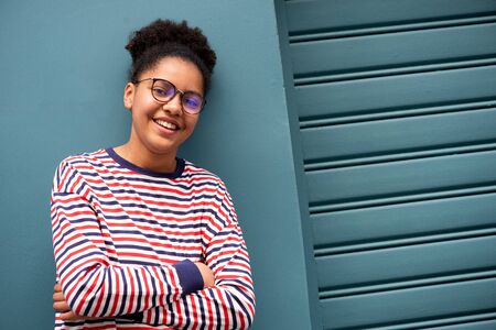 Portrait of smiling african american teenage girl with glasses and arms crossed by blue wallの写真素材