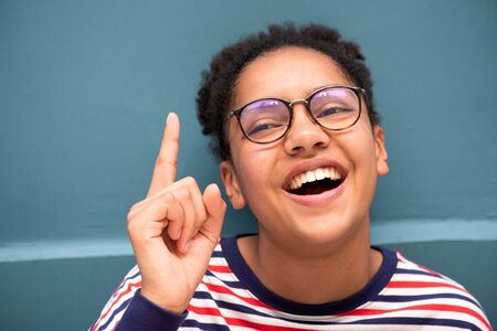 Close up portrait of smiling young mixed race girl with glasses pointing finger up against blue wall