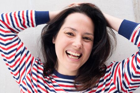 Close up portrait of happy woman laughing against white wall with hands behind head