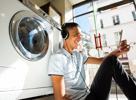Portrait smiling young man sitting on floor leaning against wash machine at laundromat listening to...