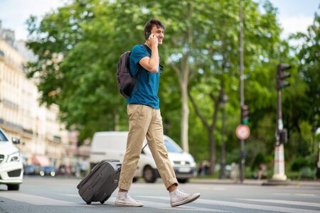 Full body side portrait of young travel man walking and talking on city street with cellphone and suitcaseの写真素材