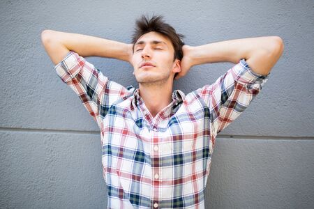 Portrait of young man leaning against gray wall with hands behind head and eyes closedの写真素材