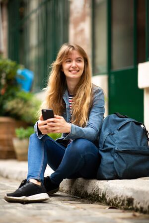 Portrait female college student sitting outside with cellphone and bagの写真素材