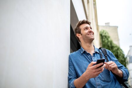 Portrait of older man leaning against wall with cellphone and looking away