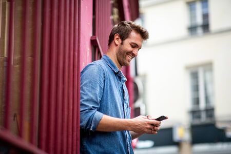 Side portrait of happy man leaning against wall looking at mobile phone in cityの写真素材
