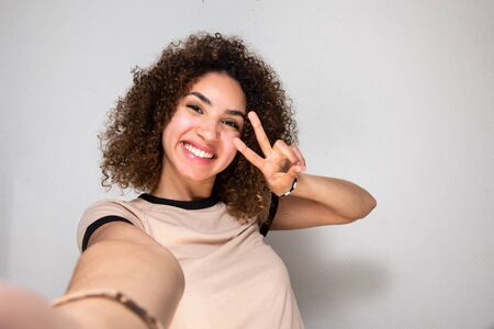 Portrait of young woman taking selfie with peace hand sign by white wallの写真素材