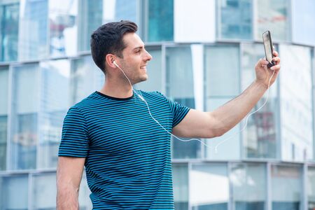 Side portrait of young man holding mobile phone to take selfie outside in city