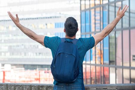Portrait from behind of young man with backpack looking at city with arms raised
