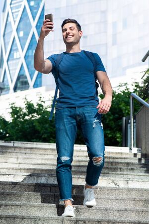 Full length portrait of happy young man walking down stairs with bag and mobile phone