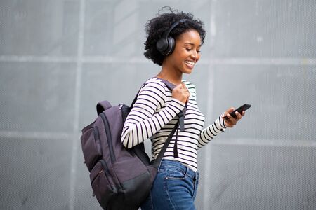 Side portrait happy young black woman traveler listening to music with cellphone and headphones and...