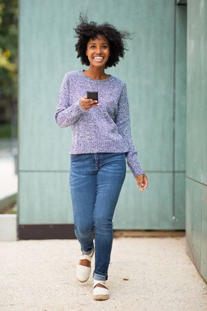 Full length portrait happy young african American woman walking outside with mobile phone