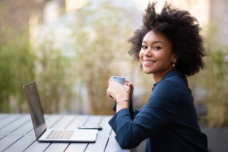 Side portrait smiling young african american business woman sitting with laptop and cup of coffee